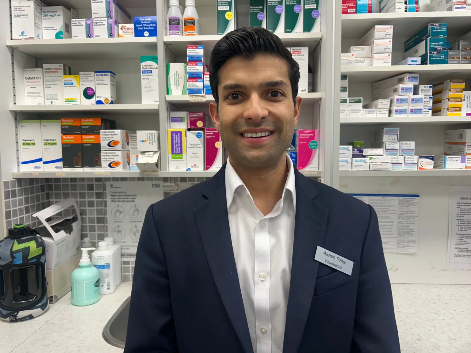 A young man in white shirt and navy blazer stands in a pharmacy smiles at the camera. 