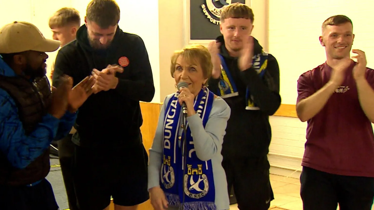 Philomena Begley is an older woman with short dark blonde hair. She is draped in a blue and white football scarf and is singing into a microphone. She is surrounded by a number of football players who are clapping along. 