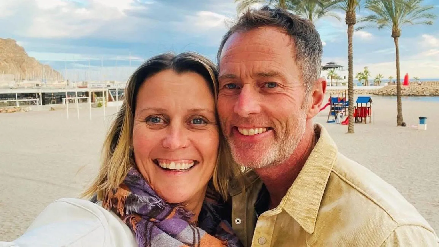 A couple on a beach. It is sunny and the sand is white. Behind them are some trees.