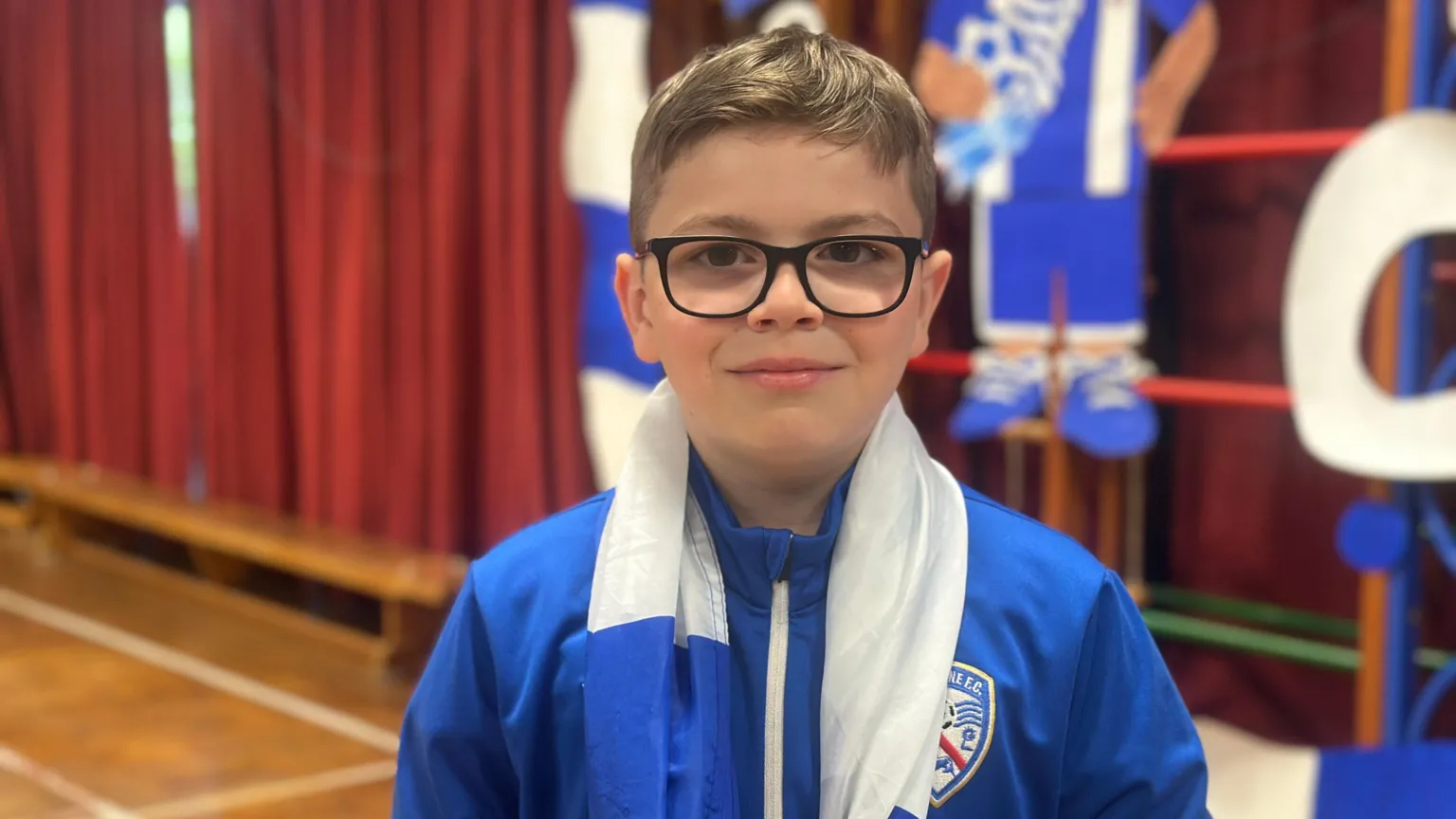Leo is a young boy of around 10 years of age. He has light brown hair worn short on the sides, and longer on the top. He is wearing dark framed glasses, a blue Coleraine FC zip up top and has a blue and white flag draped around his neck. 
