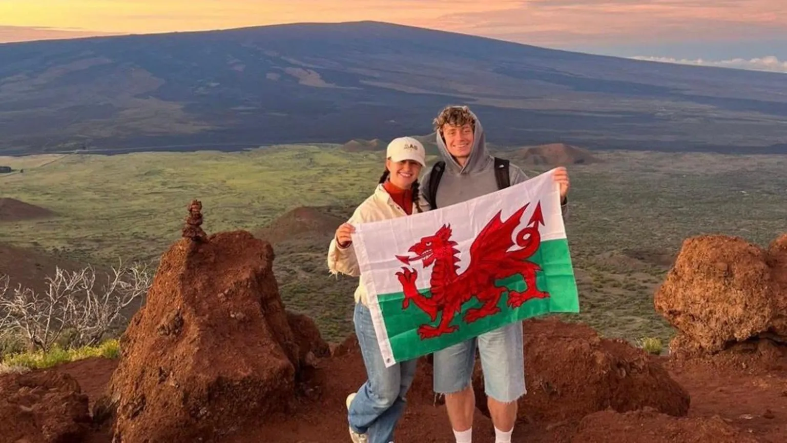 Hannah Cleaver Hannah and Jack are together holding the Wales flag. They are in front of a green field and large mountain. The sky is an orangish and yellowish colour as the sun is starting to set. 