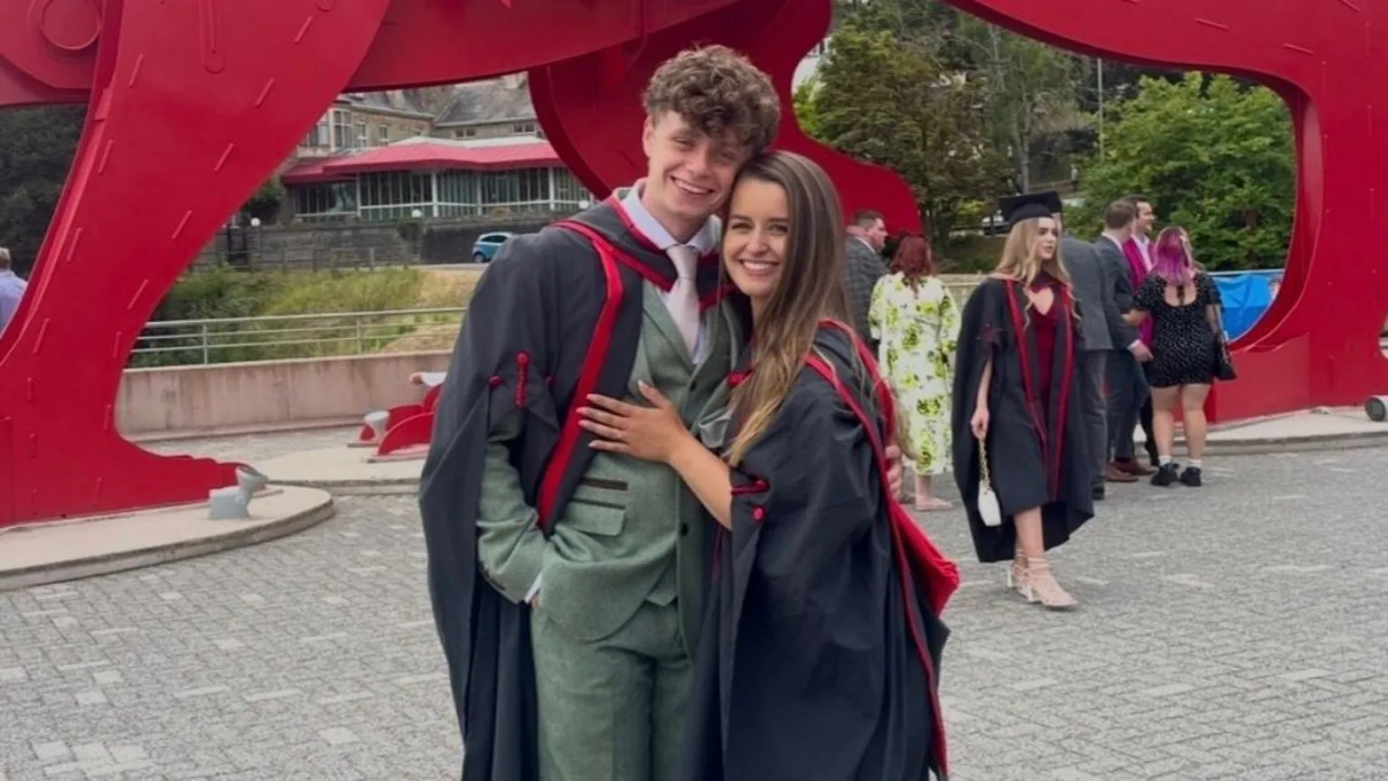Hannah Cleaver Hannah and Jack are smiling towards the camera and are next to each other. They are both wearing a red and black university gown. Jack is also wearing a light green suit. 