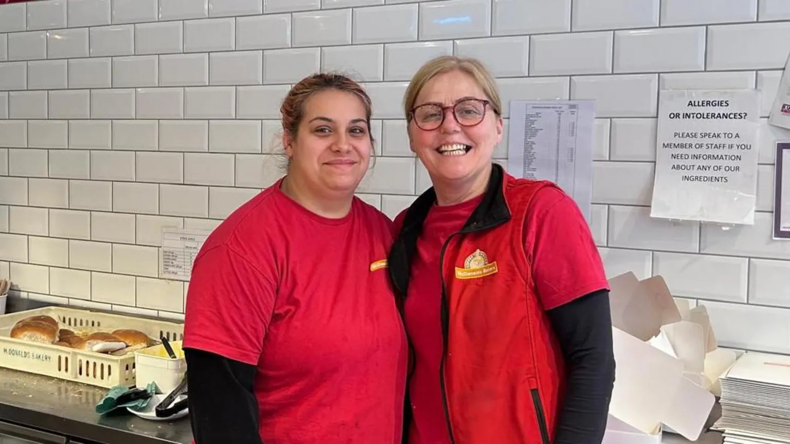 Margaret Boyd and her colleague in the bakery. Both are wearing a red uniform and smiling at the camera.