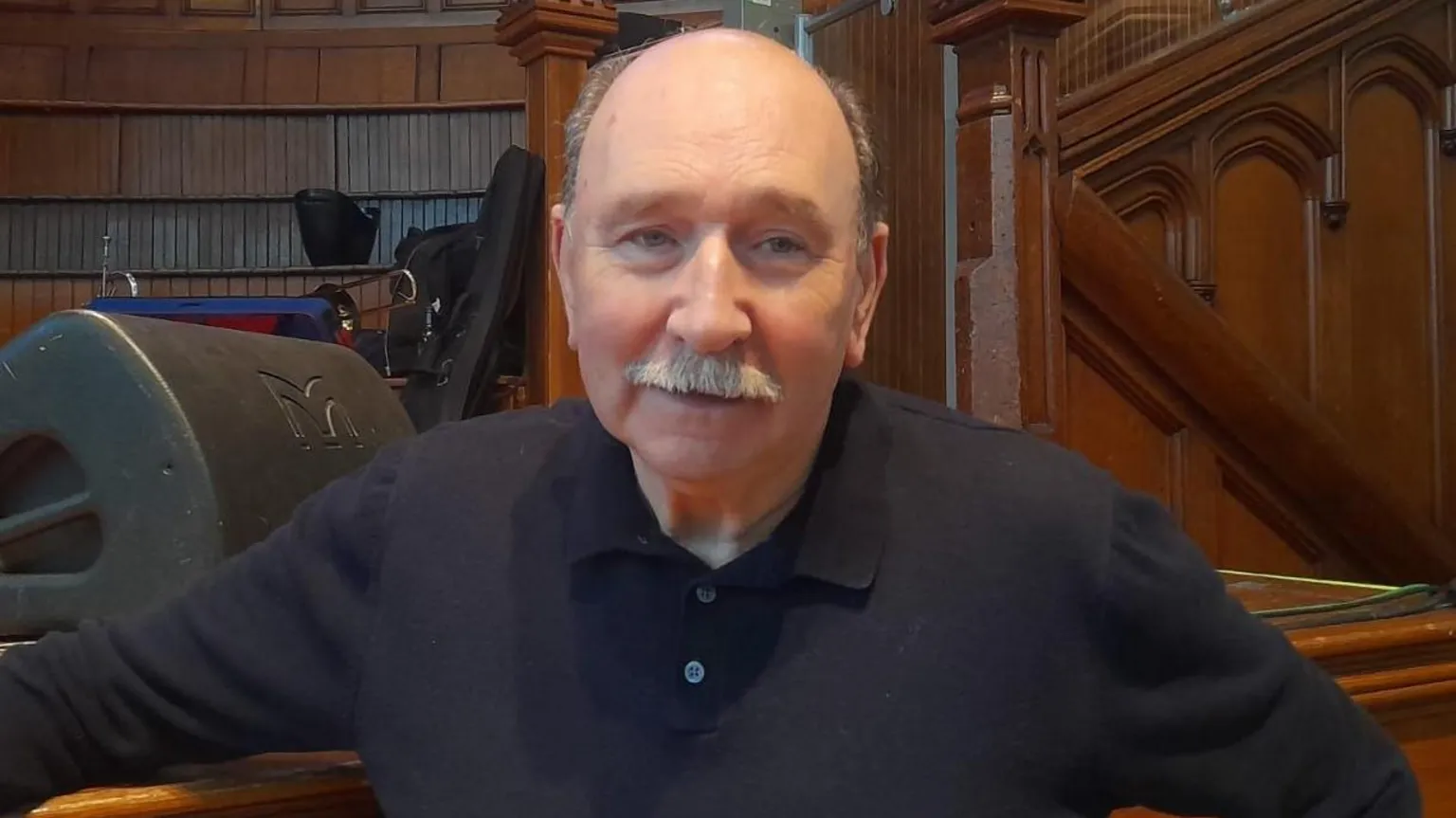 A man stands in front of a stage, wearing a black shirt, and smiling. He has a grey moustached and is balding