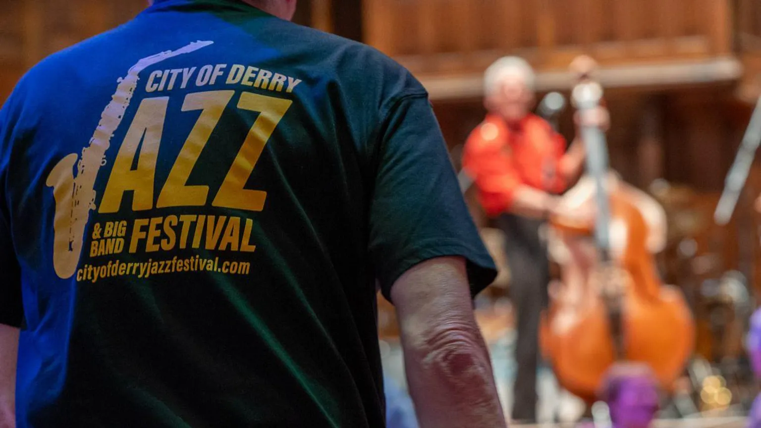 Derry City and Strabane District Council A man stands in the foreground of the image, seen from the back. He is wearing a Derry Jazz festival t shirt. In the background is a stage, where a man in a red shirt if playing a double bass