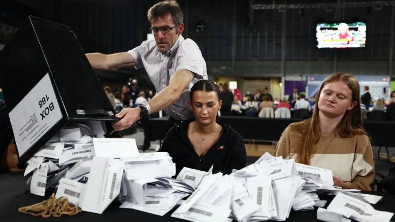  An image from the 2024 general election count in Glasgow. A man in glasses and a short-sleeved shirt is pouring a mass of ballot papers out onto a long table from a large black bin, while two younger women look on impassively, ready to start counting
