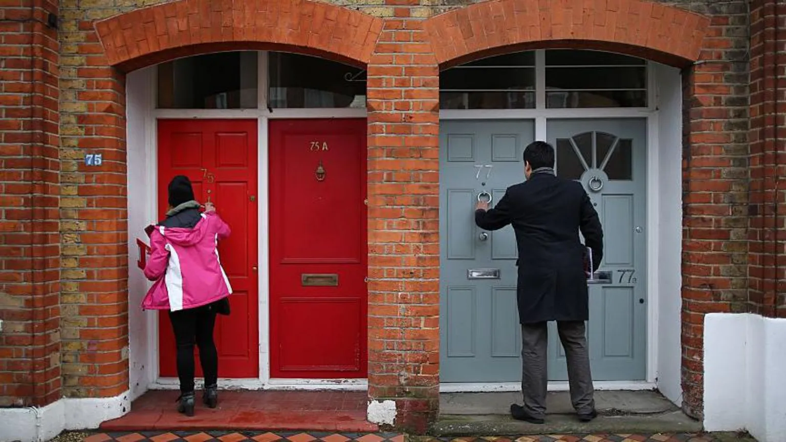  An image of two unidentified political campaigners knocking at doors - there are two red doors to the left of frame where a woman in a pink coat and black hat is standing holding a clipboard, while on the left a man in a black coat and grey trousers is reaching up to the door knocker of a grey door