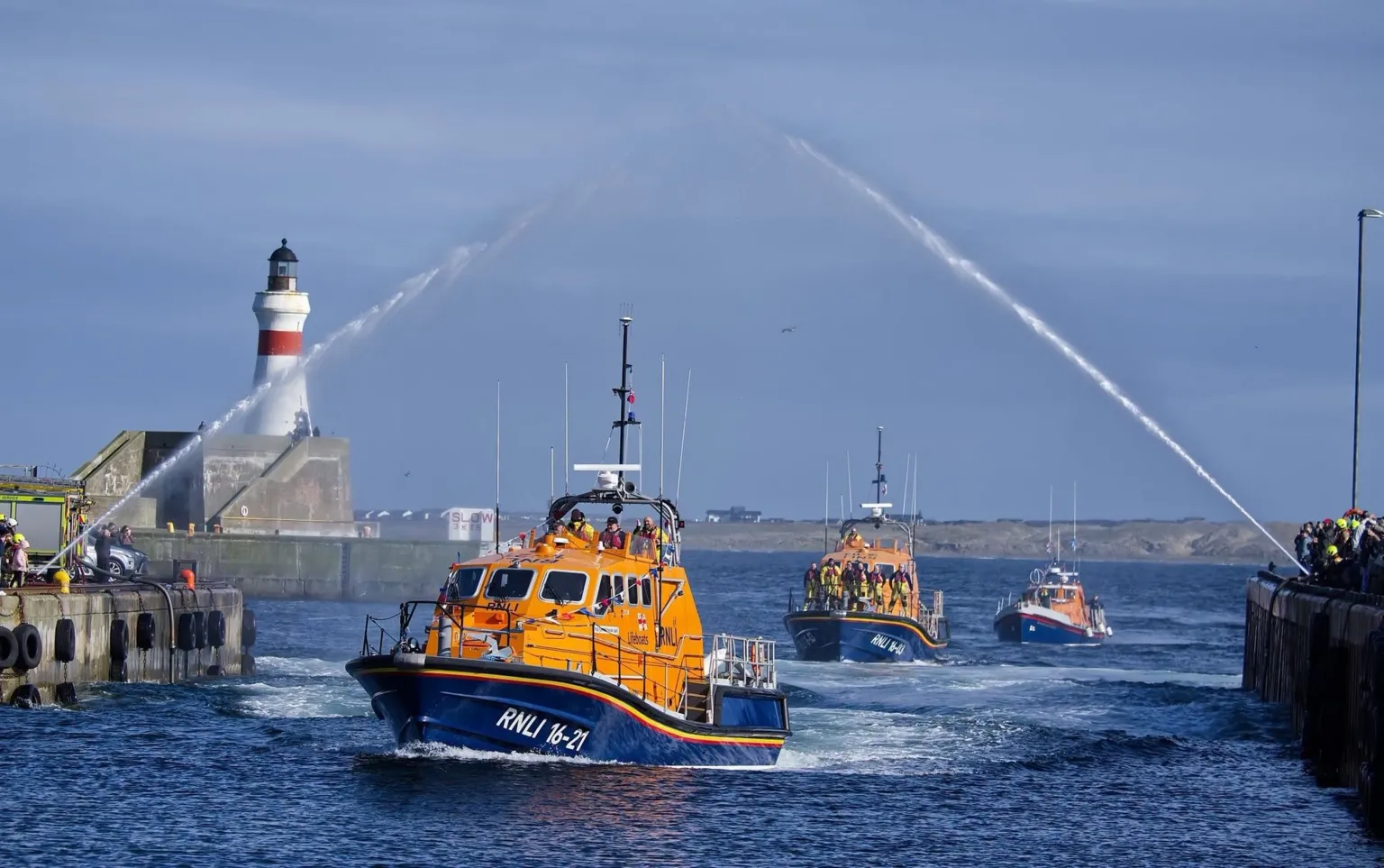 Mark Grant A parade of three orange RNLI lifeboats sailing into the harbour. There are crowds along either side of the entrance and a water arch is over the parade.