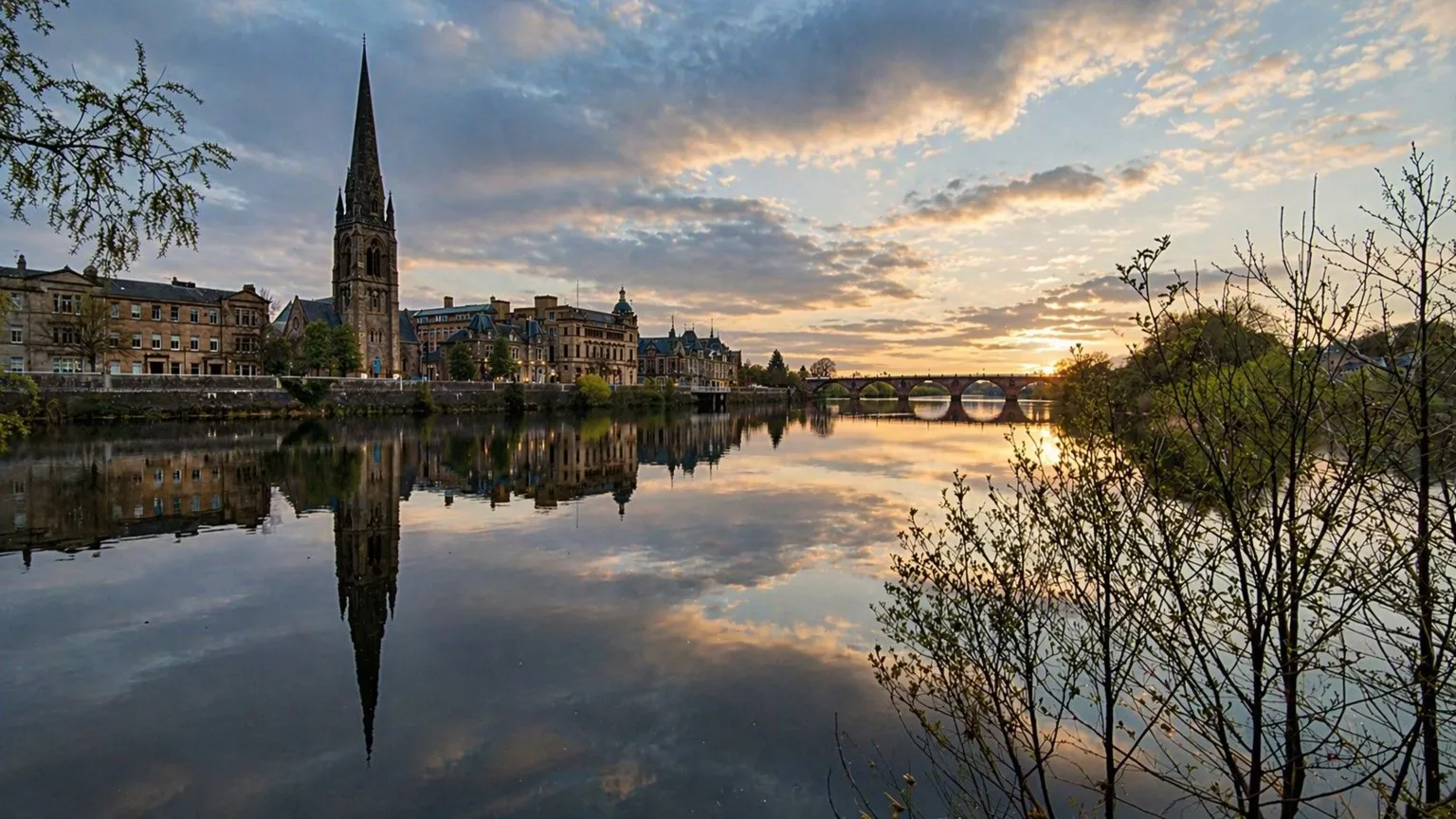 Michael O'Kane Buildings, including the spire of a church, line the river and are reflected in the water. There is a bridge in the background and the sky is yellow and blue during sunset. There are trees and bushes on the right.