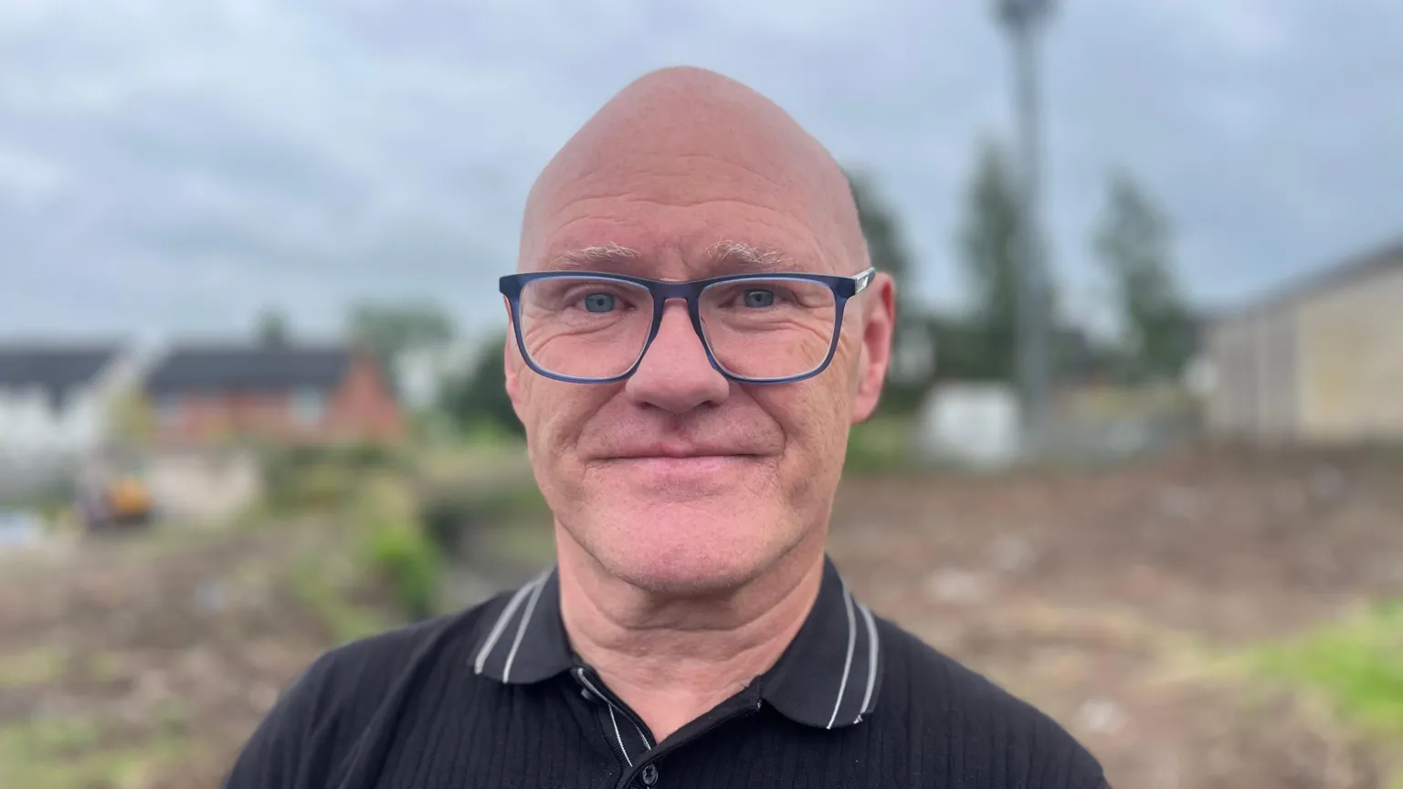 A bald man with glasses wearing a black polo shirt standing outside during the day in front of a muddy field. Houses can be seen in the background. The background is out of focus.