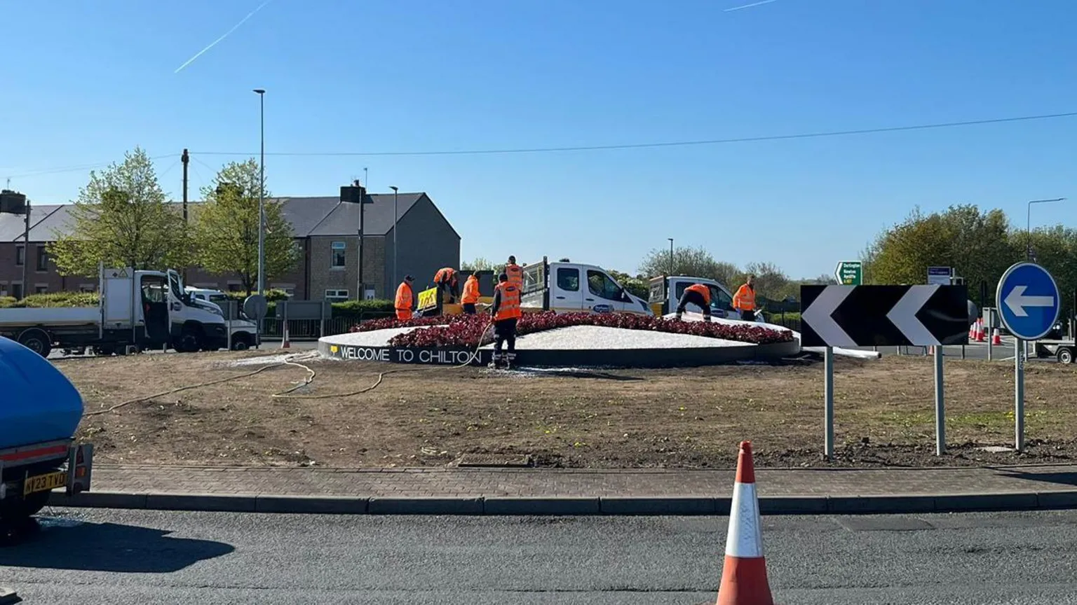 Workmen in orange hi-vis vests are pouring white gravel and planting red flowers into a raised bed in the middle of the roundabout. It is circular and taking the pattern of the St George's cross. Traffic cones have been placed around the inner lane of the roundabout.