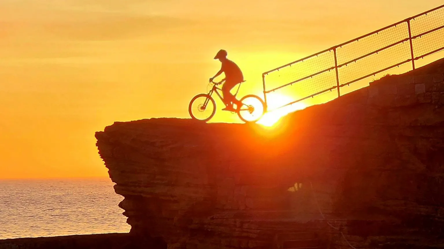 Rhona Graham Silhouette of a cyclist riding along a cliff edge at sunset, with the sun glowing behind the bike and the sea in the background.