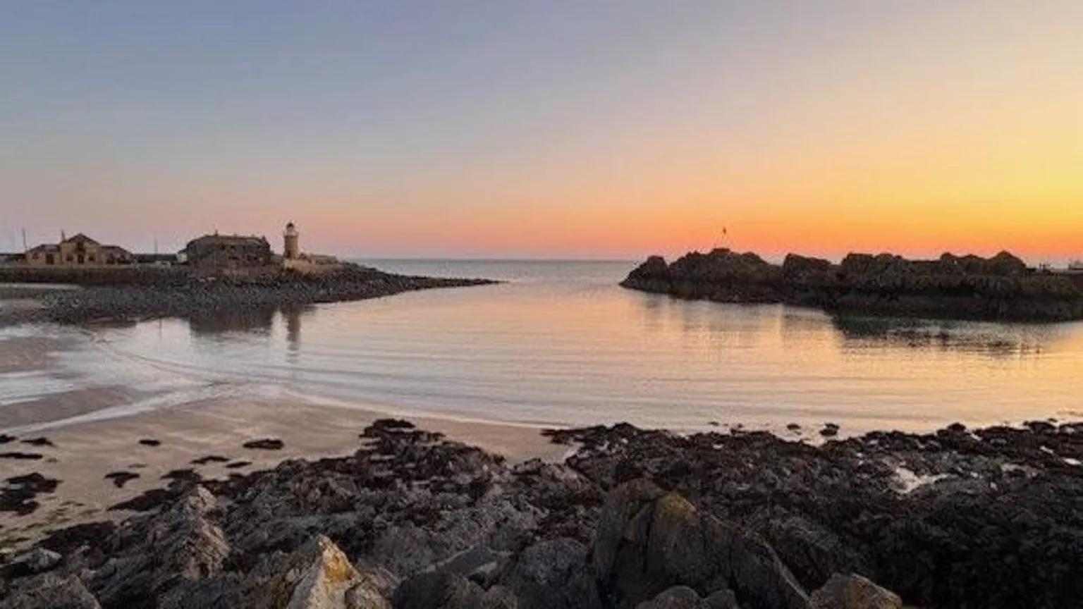 Karen Ritchie Wide view of a calm coastal harbour at dusk, with rocky shore in the foreground, still water reflecting pastel sky, and a small lighthouse and buildings on the horizon.