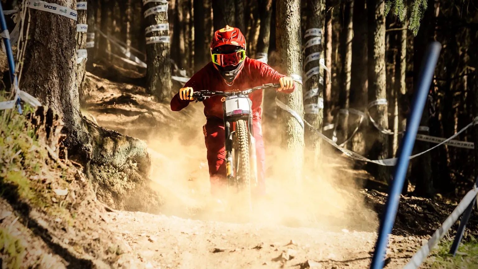 Keith Valentine A Mountain biker wearing red overalls and a red helmet racing downhill through a forest trail, kicking up dust between trees.