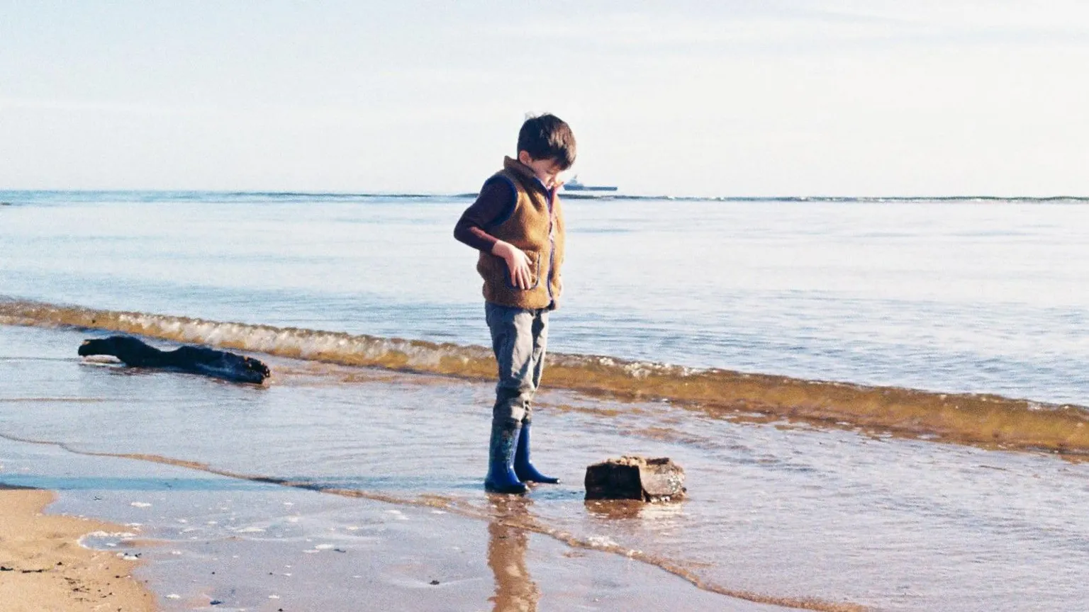 Ian Adamson A Young boy standing at the shoreline, looking down at a rock as gentle waves wash over wet sand on a calm, bright day. He is wearing blue wellies and a tweed bodywarmer.