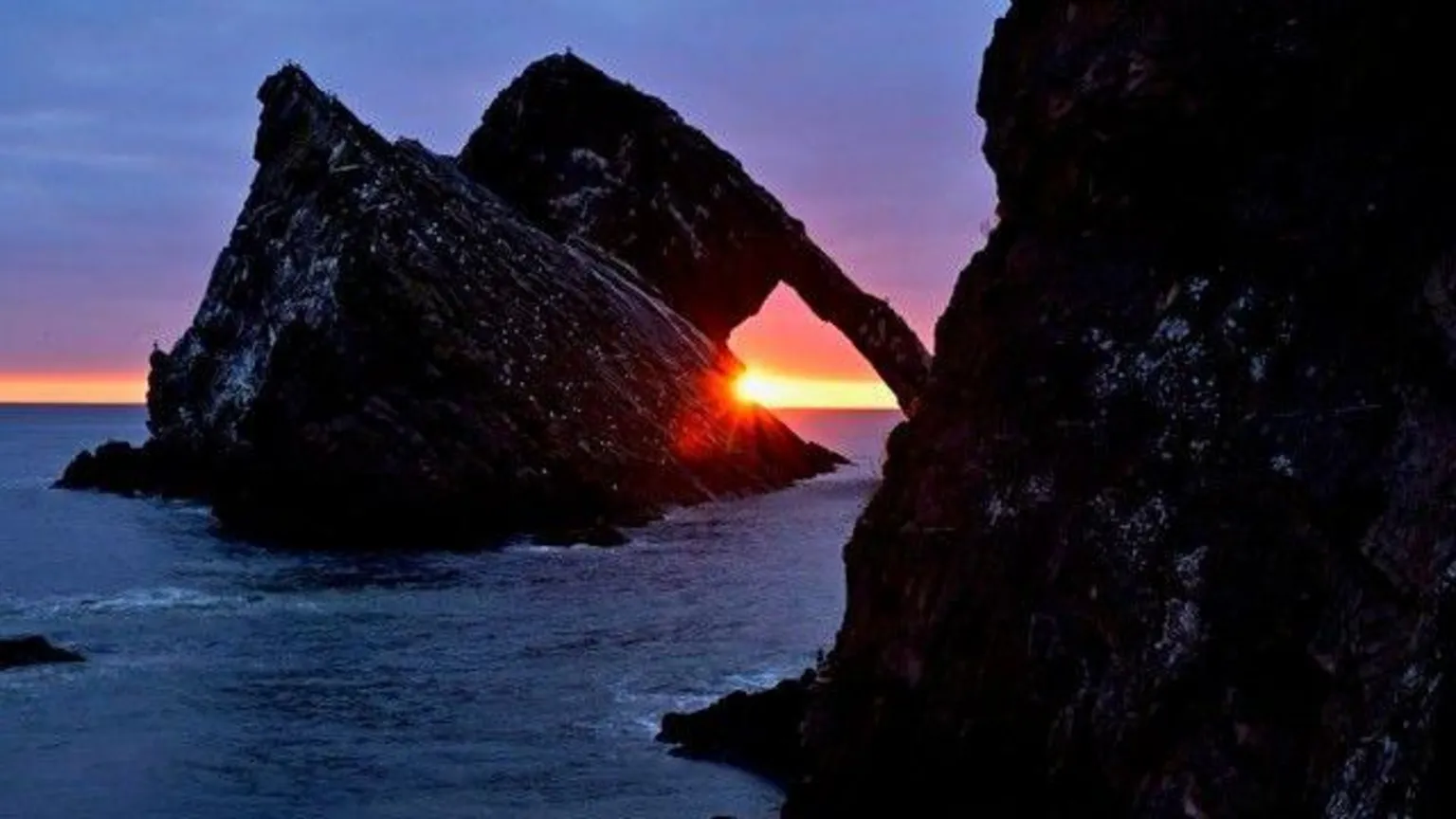 Stephen Dixon Sun setting through a natural rock arch over the sea, with dark cliffs silhouetted against a deep orange and purple sky.