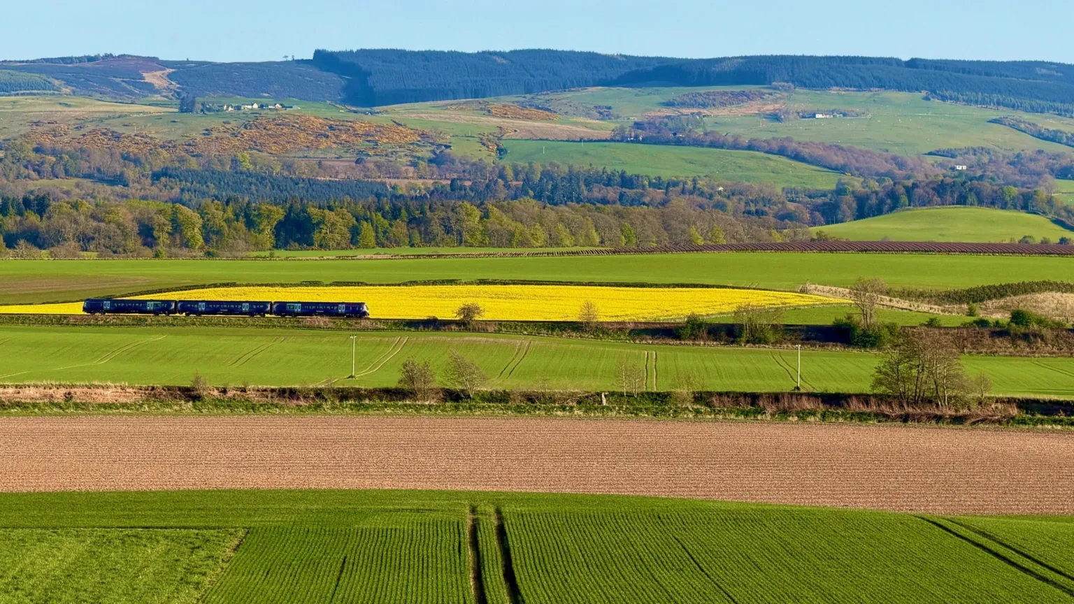Lorna Donaldson A Passenger train travelling through patchwork farmland, with green and yellow fields stretching toward wooded hills under a clear blue sky.