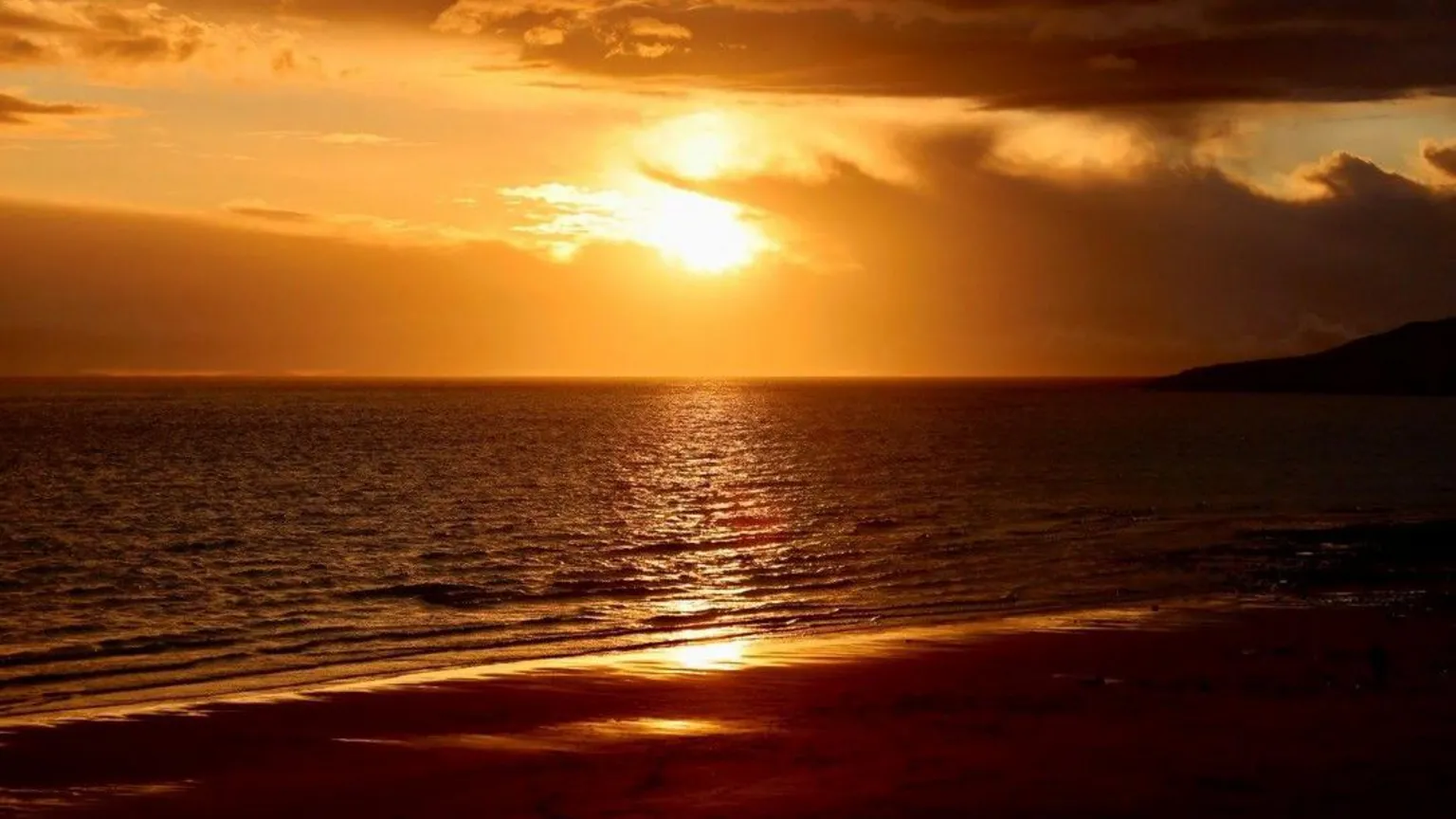 Graham Christie a golden sunset over a calm sea, with dark clouds drifting across the sky and warm light reflecting on gentle waves along the shoreline.