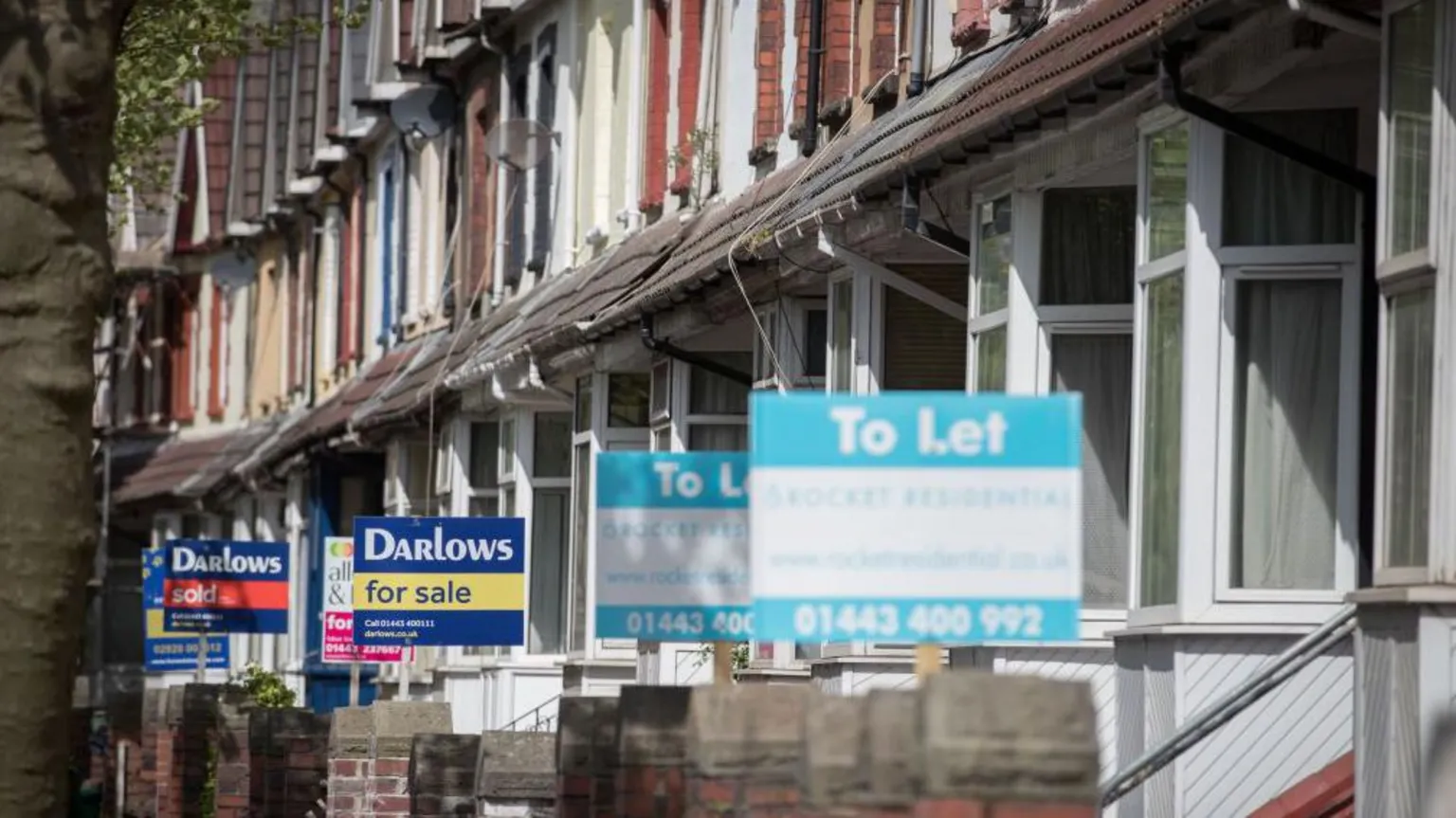 The photo shows a row of houses with bay windows and different colour bricks. Most have a to let or for sale sign showing. The front sign is slightly blurred with the signs further down the street more in focus.