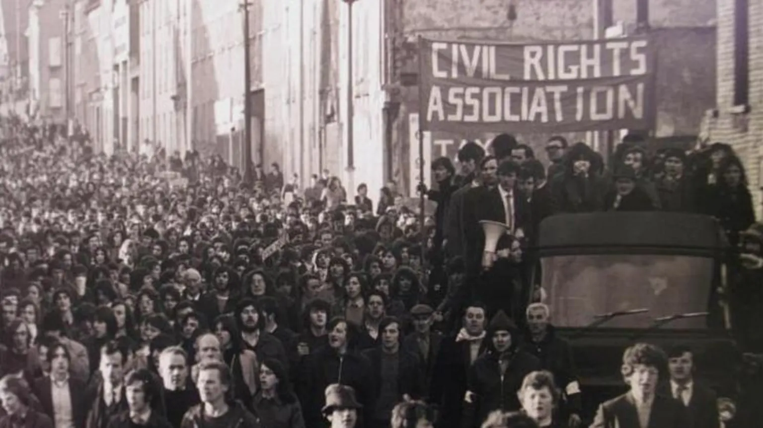  A black and white image of crowds gathered. People on the back of a truck are holding up a banner reading: 