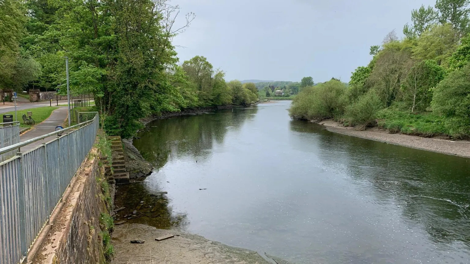 A winding stretch of the River Nith with gravelly banks and trees down either side with hills in the distance and hazy, cloudy, light blue sky overhead. There is a footpath on the left hand side of the river, and steps can be seen going down into the water. 