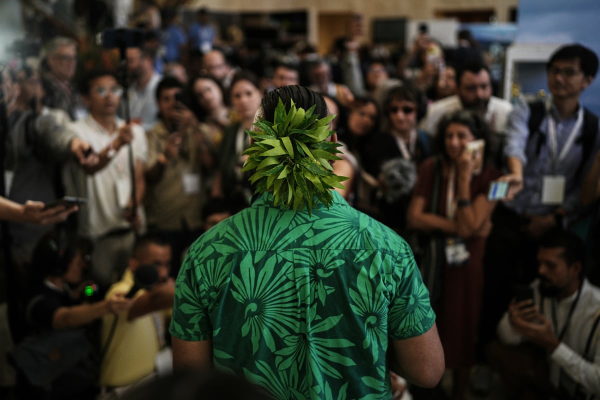 Back of Sikulu wearing green shrt and headpiece as he faces audience