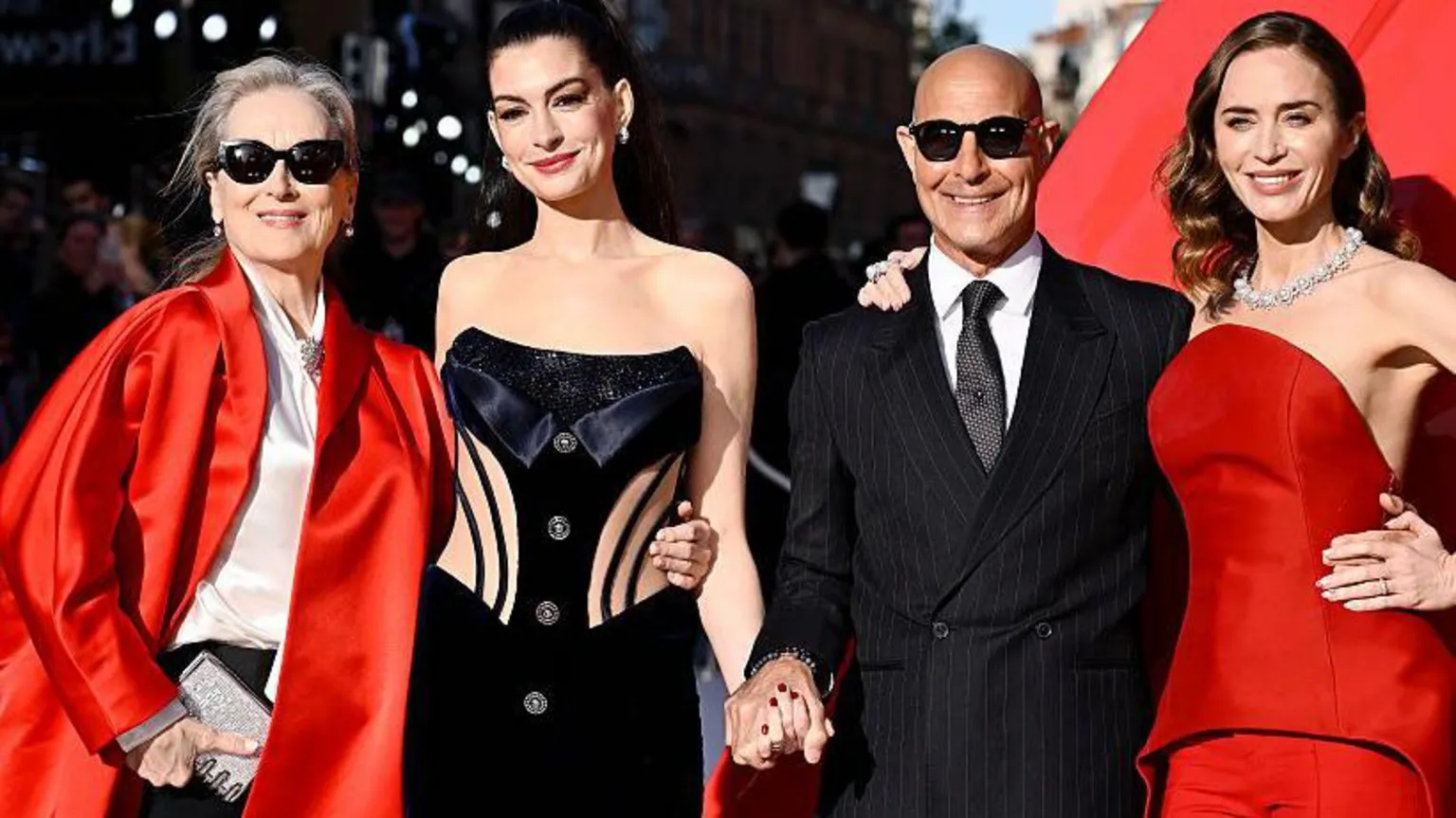  From left to right: Meryl Streep, Anne Hathaway, Stanley Tucci and Emily Blunt. The group of four stand on the red carpet at the film's premiere. Meryl wears a bright red jacket with a silky top underneath and is wearing sunglasses. Anne wears a cut-out black dress with a bow on the front and has her hair up. Stanley wears sunglasses with a pinstripe suit and Emily wears a sleevless dark red top and trousers with a bright silver necklace 