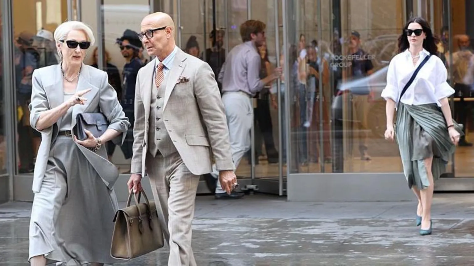  On the left, Meryl Streep is seen wearing a light grey blazer and skirt with a brown belt and dark sunglasses. Her hair is in a quiff and Stanley Tucci is next to her in a beige suit as they walk away from Anne Hathaway's character in the background