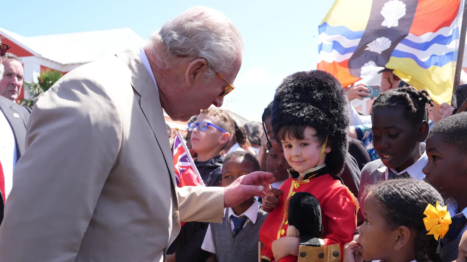  King Charles III speaks to a little boy in a guardsman outfit. The little boy is wearing a teddy bear that also wears the outfit.