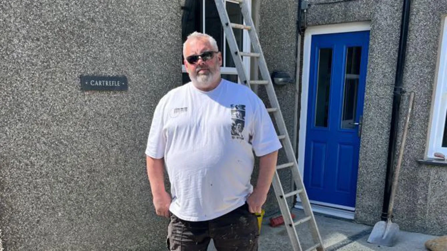 Jason is wearing a white t-shirt and black shorts. He has grey hair and beard and is wearing black sunglasses. He is stood in front of a pebble-dashed house. It has a blue door and a ladder and shovel in view behind him.