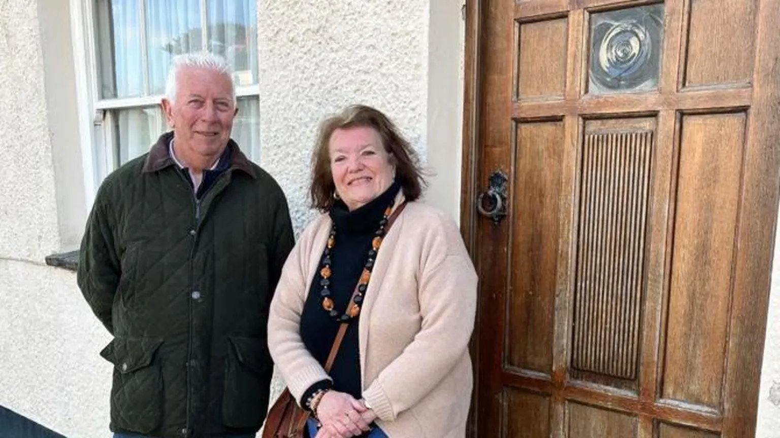 Stephen is stood on the left wearing a black quilted jacket. His hands are folded behind his back. He has white hair. Jean is stood on the right. she has her hands folded in front of her. She wears a black polo neck jumper, a cream cardigan and has a brown handbag over her shoulder. They are stood in front of a house with a brown door.