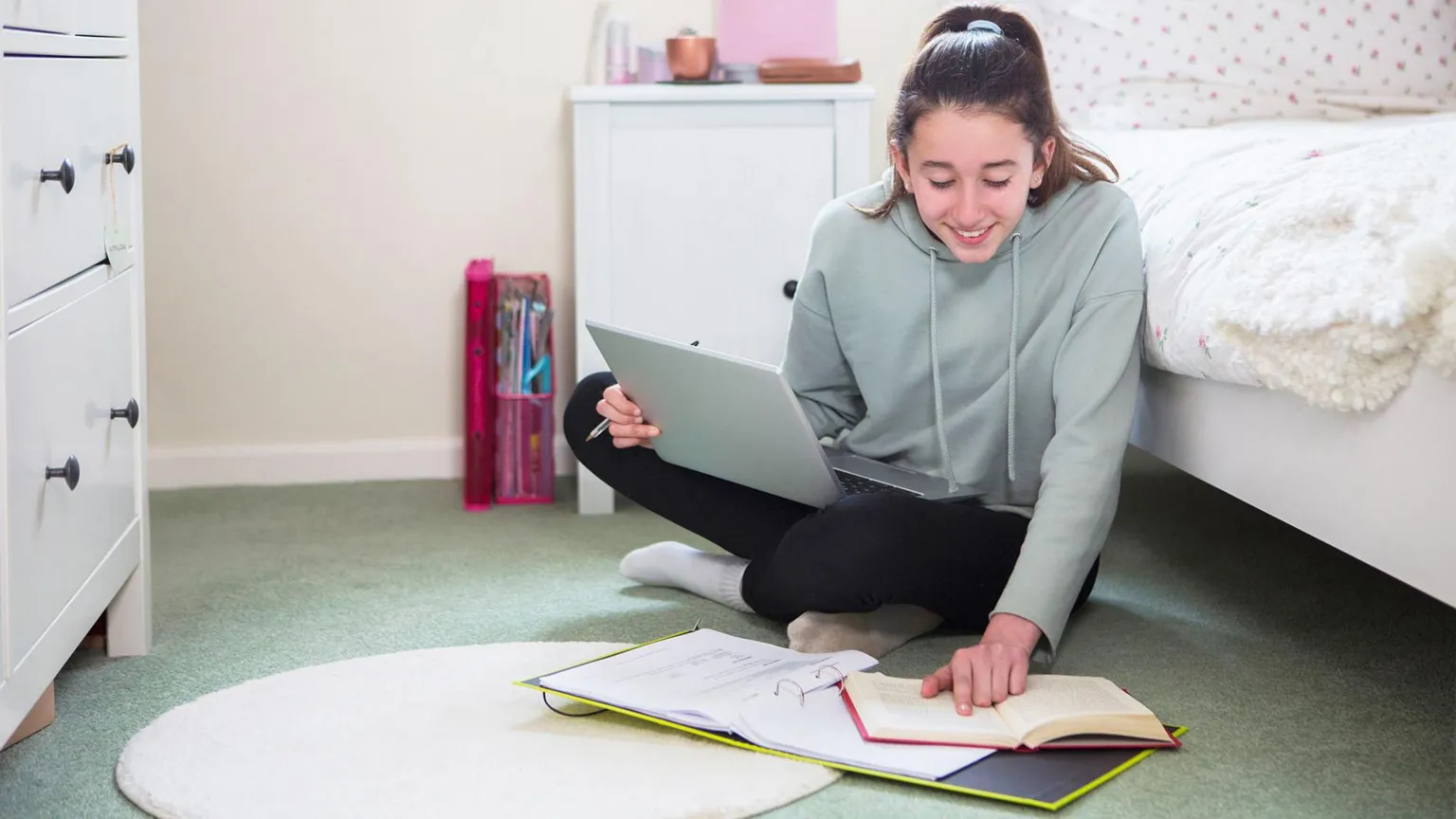  A girl wearing a light green jumper with brown hair tied up in a ponytail, she is sat on her bedroom floor with her laptop on her legs and her folder and notebook on the floor, she is smiling and is reading her notes. 