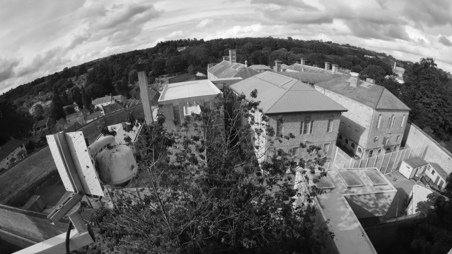 Dave Cable A black and white aerial view of Shepton Mallet prison, there are several large block buildings surrounded by walls and trees in the foreground. 