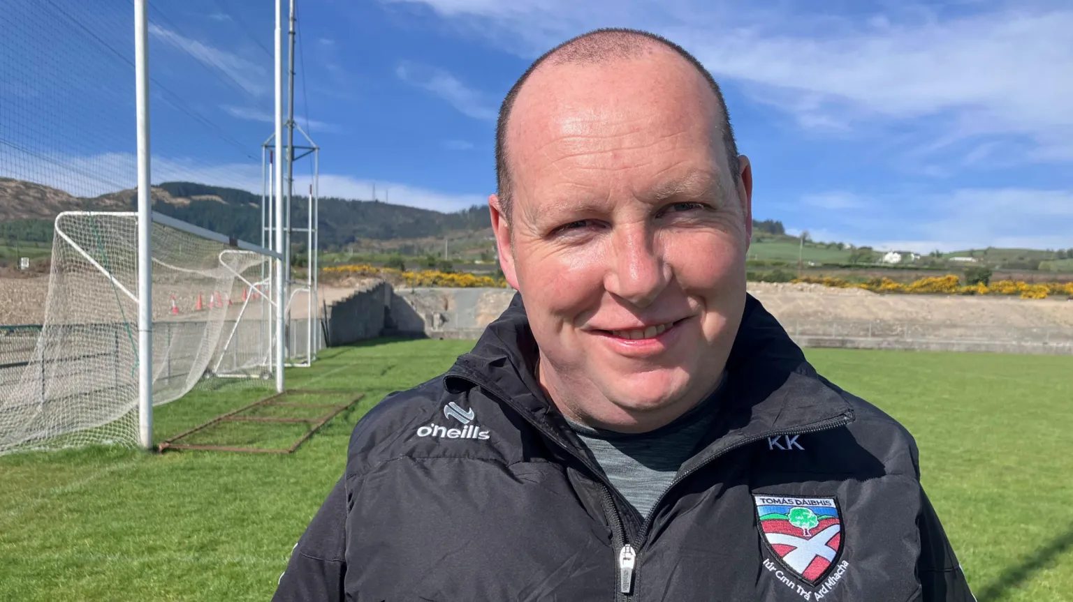 Karl Simmons has tightly shaved dark hair. He's dressed in a black waterproof O'Neill's sports jacket with a badge on the top. Behind him is a gaelic football pitch and white posts. 
