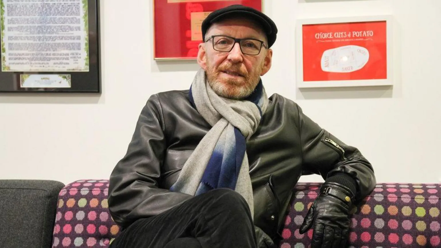 A man with a reddish-grey beard wearing glasses with a grey and blue scarf on, flat black cap and leather jacket. He is sitting on a spotted sofa.