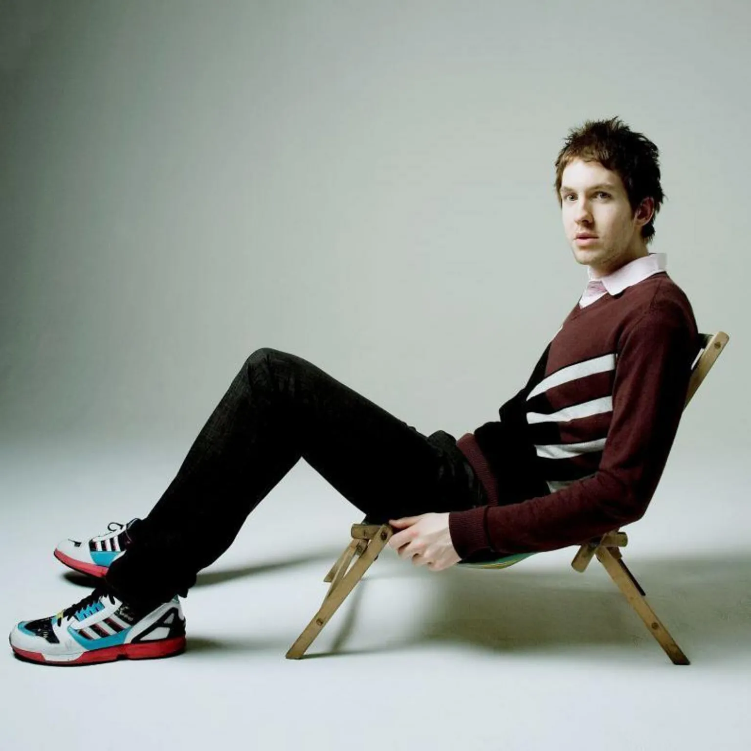  Calvin Harris with very dark hair. He is sitting in a low deckchair, wearing black jeans, trainers and a brown jumper. 