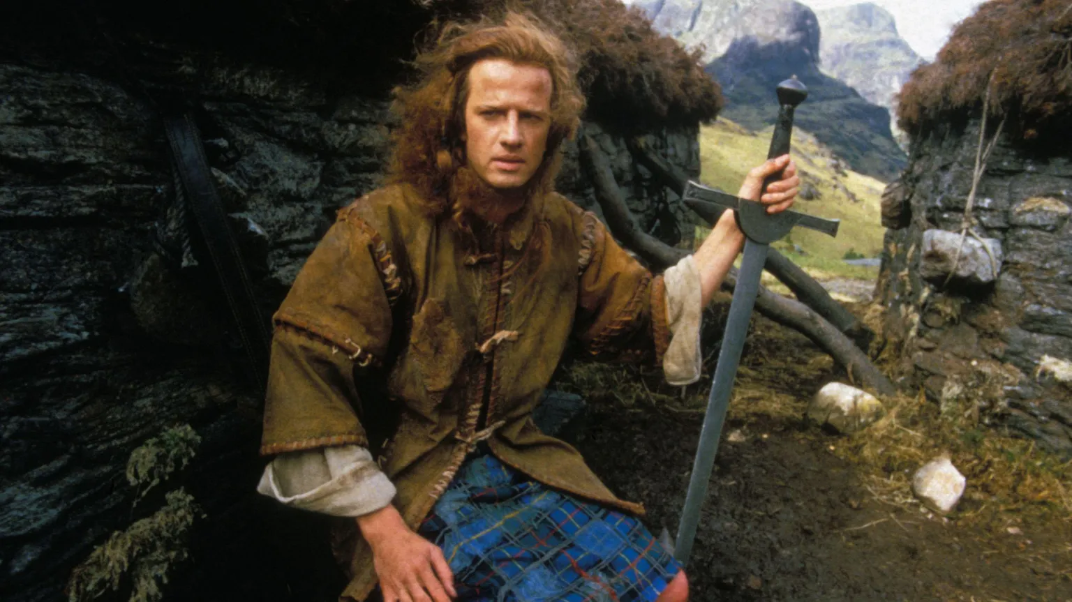 Georges De Keerle/Gamma-Rapho via Christopher Lambert is wearing period Highland garb and is holding a sword as sits by the wall of a stone walled, turf roofed building. A glen and mountains are behind him.