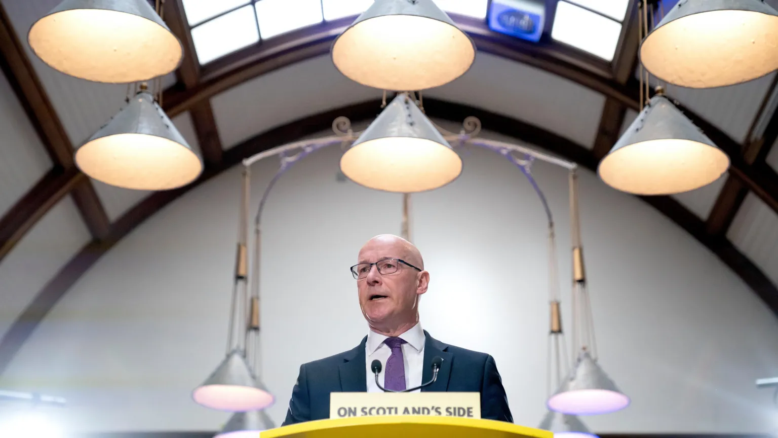  John Swinney, who is bald and wearing glasses, is standing behind a lectern in a domed room with lights hanging above his head. He is wearing a blue or grey suit, white shirt and purple tie. There is a sign in front of him that says On Scotland's Side 