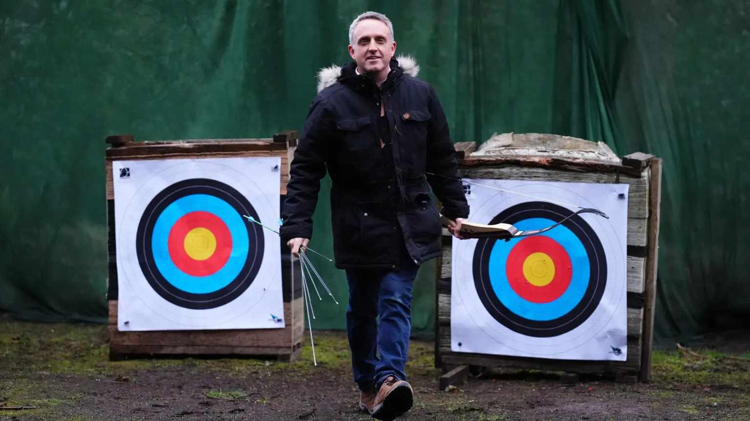  Alex Cole-Hamilton walks towards the camera, holding a bow in his left hand and arrows in his right hand. There are two large targets behind him and he is wearing a dark blue jacket with a furry hood and blue jeans.