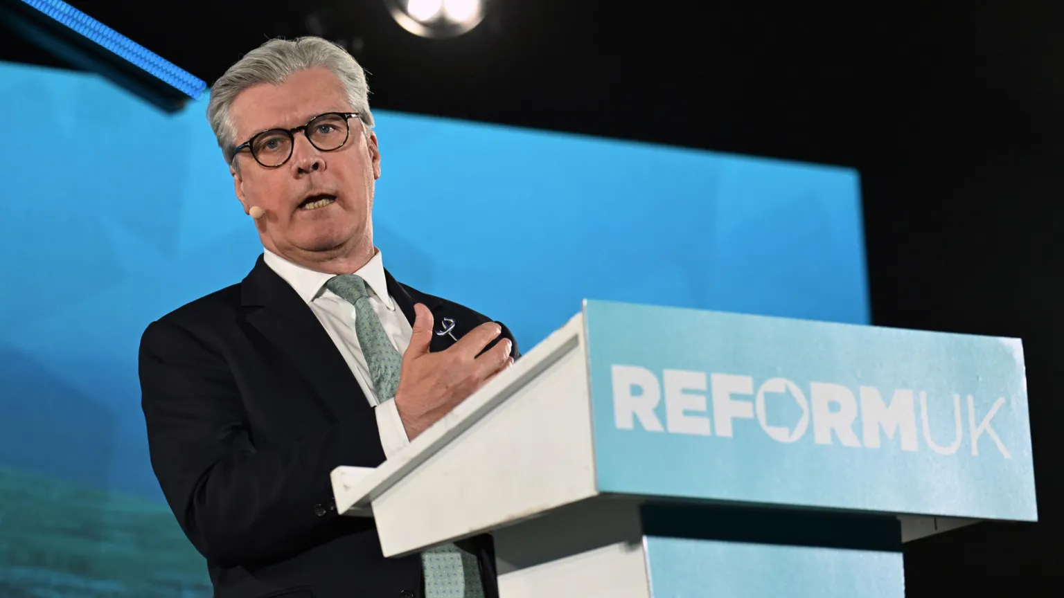  Malcolm Offord standing on stage behind a lectern with Reform UK written on it. He has grey hair and glasses and is wearing a dark suit, white shirt and green tie