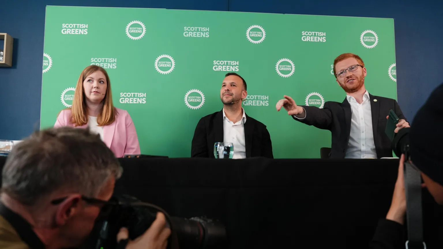  Gillian Mackay, Zack Polanski and Ross Greer sitting behind a table at a press conference, with a Scottish Greens backdrop behind them.