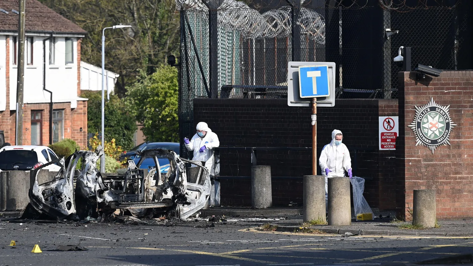 Charles McQuillan/ Two forensic investigators at the scene of the Dunmurry car explosion. They are both wearing white hazmat suits, blue masks and purple plastic gloves. They are walking around the wreckage of the exploded car which is lying on the road beside concrete bollards outside the large police station wall. On investigator is carrying a yellow evidence marker inside a clear plastic bag. 