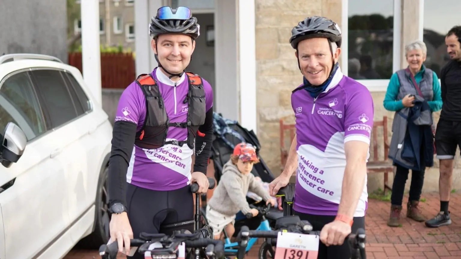 Family Picture On the right is a man stood next to a bike whilst smiling at the camera. He has dark eyes and is wearing a bicycle helmet on his head with sunglasses resting on the helmet. He has a purple t-shirt on with a black long sleeve top underneath and black shorts. Next to him is a man with grey hair who's also smiling at the camera. He's holding a bike too. He's wearing a purple t-shirt and black shorts.