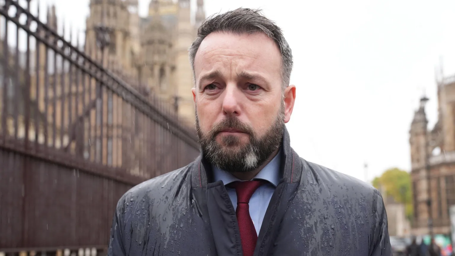  A man with black and grey hair, and beard, is standing outside looking to the left of the camera. He is wearing a coat, wet from rain, a maroon tie and light blue shirt. A fence can be seen behind him as can an old tall building. 