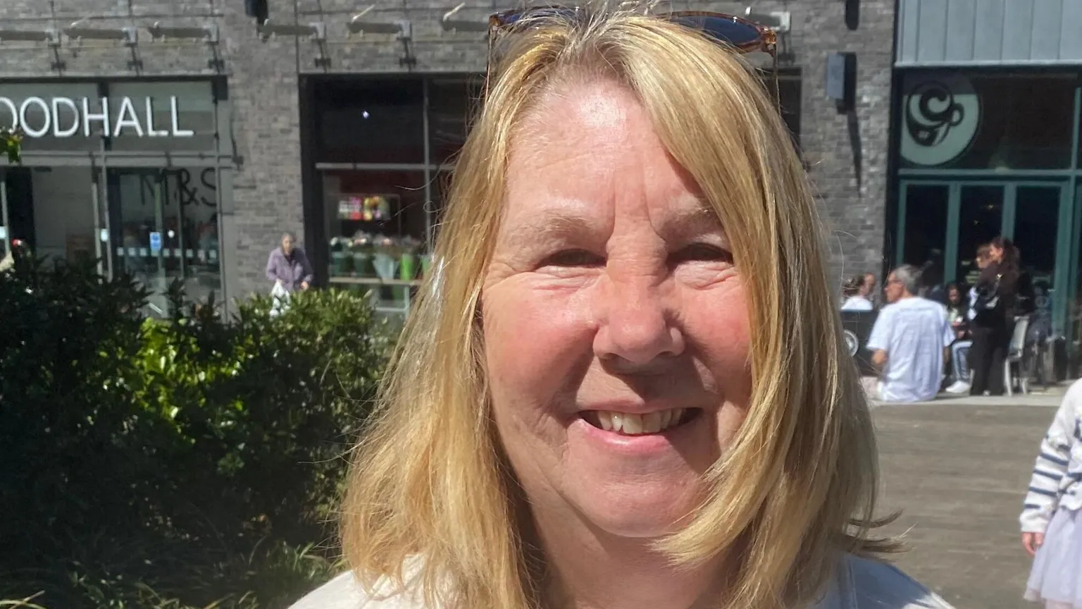 Liz Thorne, looking at the camera. She has blonde hair and is in front of a set of shops including a cafe and an M&S.