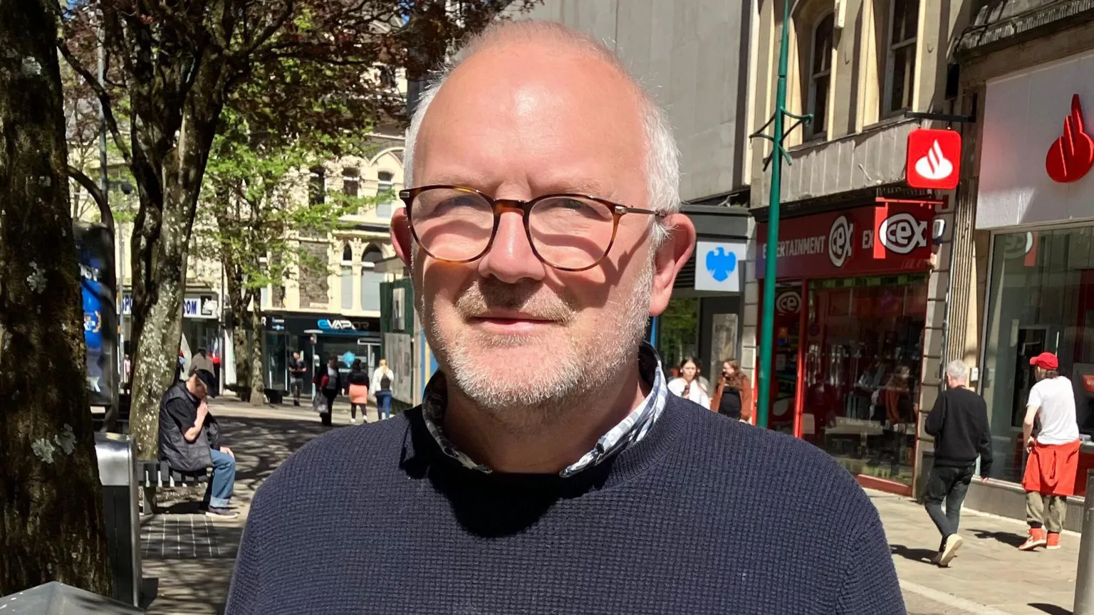 Andrew Lightbown is wearing a dark blue jumper and is looking at the camera. His arms and upper body are visible. People can be seen walking in the background in front of the city's shops.