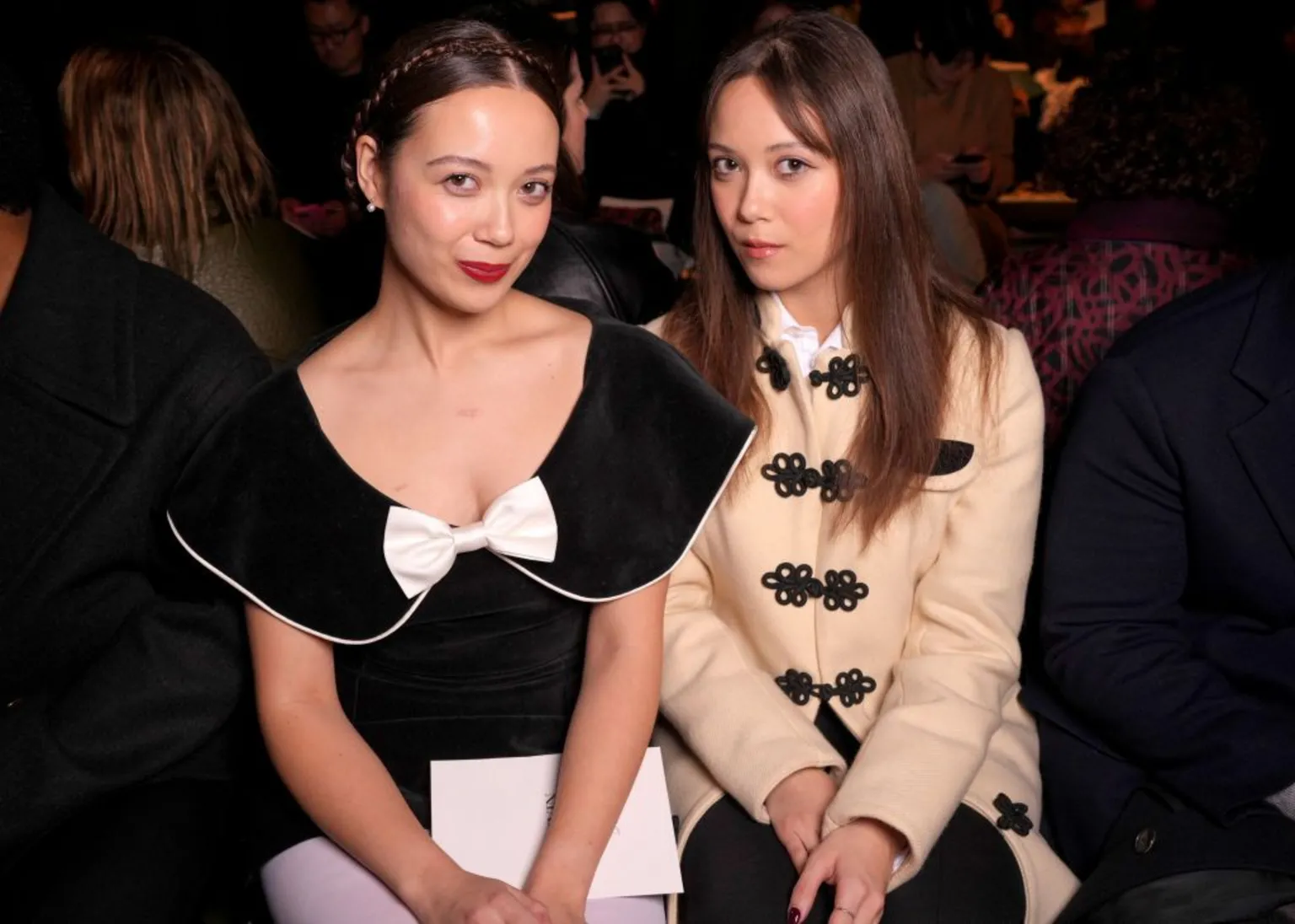  Laufey and her sister Junia sit side by side during a public event. Laufey is dressed in an elegant black gown with a white bow on the neckline; while Junia is in a tan button-up jacket, with ornate black clasps
