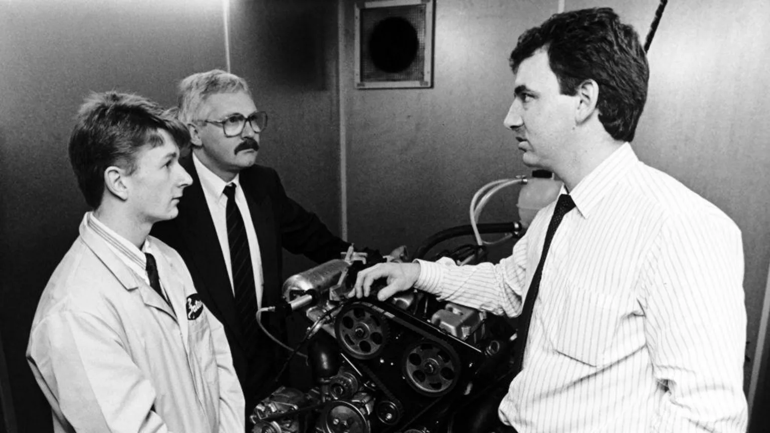  A black and white picture of a young Allan McNish in overalls talking to two other men about an engine