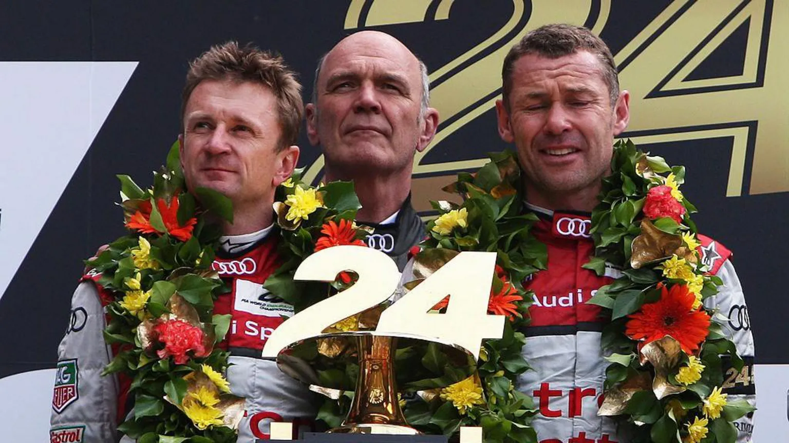  Allan McNish on the left of the podium with his two teammates after winning Le Man. They are decked in flowers and are holding a trophy with the number 24 on it.