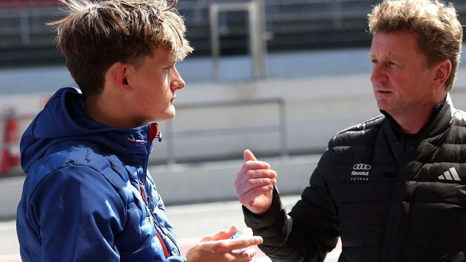  Allan McNish in a dark Audi jacket is deep in conversation with young racer Freddie Slater in a blue jacket with light brown hair. They are at a race track.
